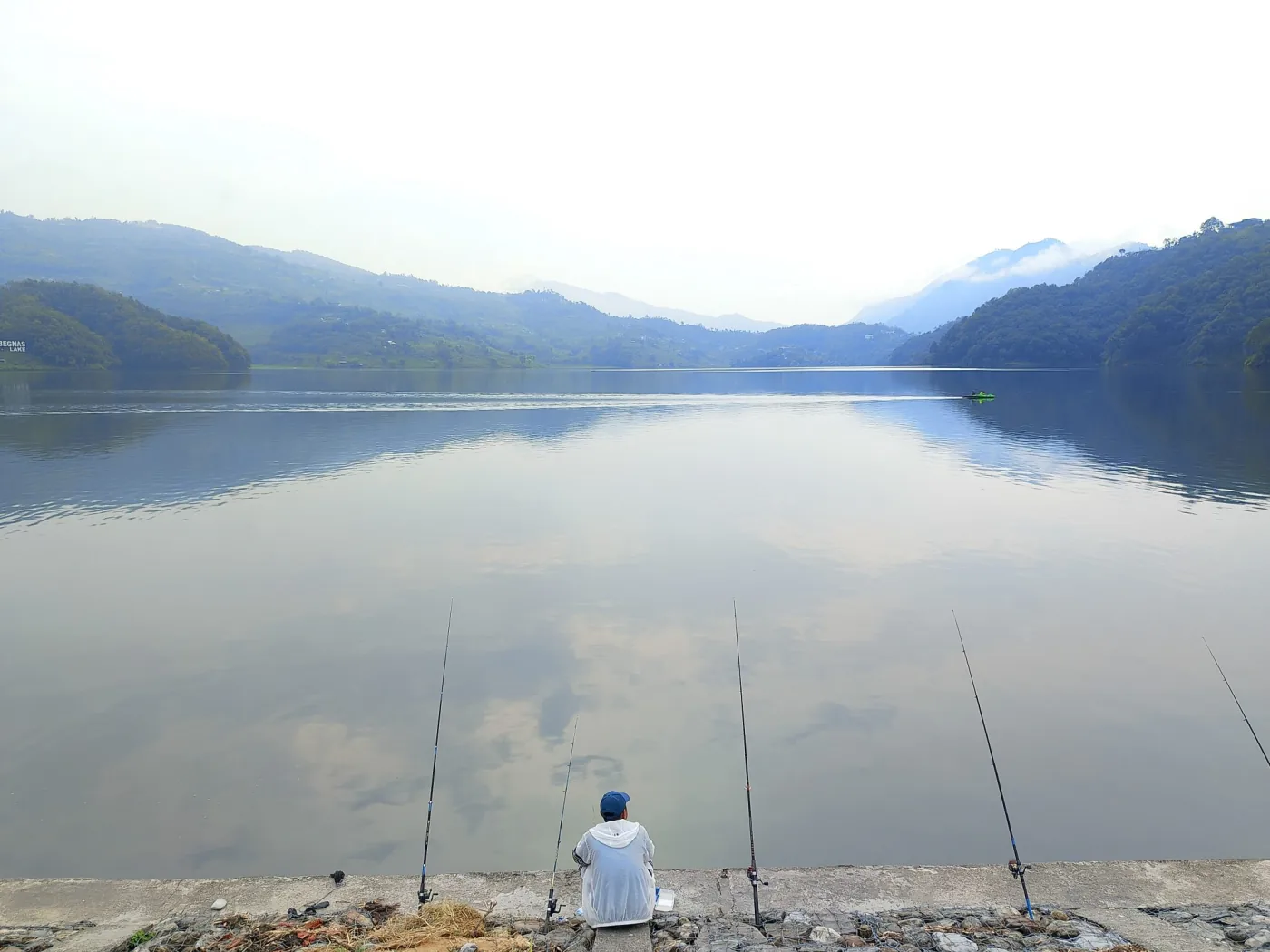 Calm lake reflecting clouds and misty green hills, with a person seen from behind sitting on a stone ledge